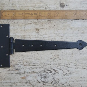 Può includere: Una cerniera nera a forma di T con un'estremità decorativa a punta. La cerniera in metallo ha diversi fori per le viti. Un righello di legno è posto sopra la cerniera, che mostra le misure in centimetri.