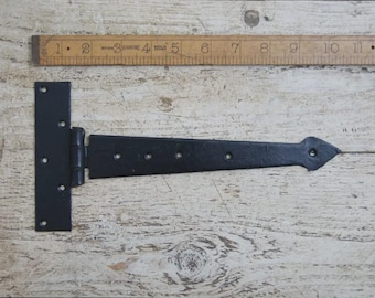 May include: A black, T-shaped hinge with a decorative pointed end. The hinge is made of metal and has several screw holes. A wooden ruler is placed above the hinge, showing measurements in inches.