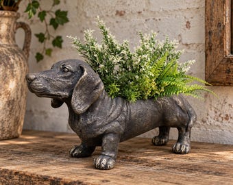 May include: A dark gray, textured dachshund-shaped planter with a weathered finish. The dog figure stands on four short legs, with a hollow back containing a cluster of green and white artificial foliage, including ferns and small flowers.