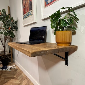 May include: A rustic wooden shelf with black metal brackets mounted on a white wall. A laptop computer is on the shelf, and a potted plant with green leaves is on the shelf to the right of the laptop. The plant is in a yellow striped pot. There are two framed posters on the wall behind the shelf.