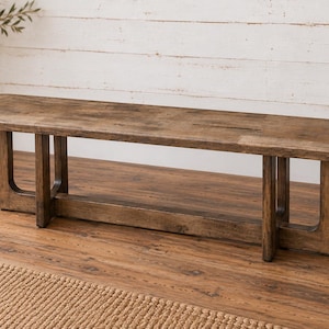 May include: A rectangular wooden bench with a medium brown finish and visible wood grain. The bench features a flat seat with rounded corners and is supported by four vertical legs connected by a horizontal base stretcher on each side.