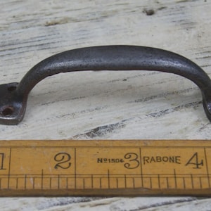 Könnte beinhalten: Ein dunkelgrauer, gebogener Metallgriff mit Montageplatten an jedem Ende. Der Griff ist etwa 10 cm lang und hat ein rustikales, gealtertes Aussehen. Unter dem Griff liegt ein Holzlineal.