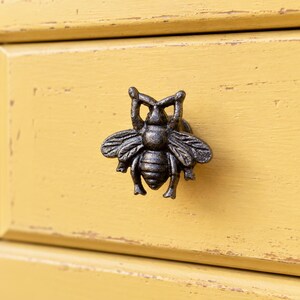 Puede incluir: Un pomo de cajón de metal oscuro con forma de abeja y acabado texturizado. La figura del insecto presenta alas detalladas, un cuerpo segmentado y antenas elevadas. Está montado sobre una superficie de madera pintada de color amarillo.