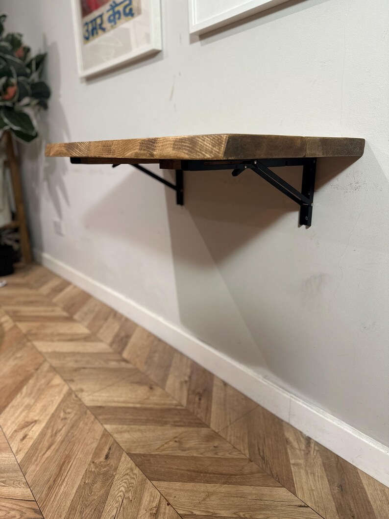 K&ouml;nnte beinhalten: Ein an der Wand montierter Klapptisch aus Holz mit schwarzen Metallkonsolen. Der Tisch ist aus recyceltem Holz gefertigt und hat eine rustikale Oberfl&auml;che.
