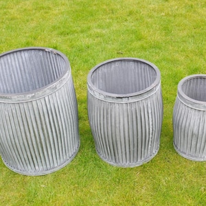 May include: Three galvanised metal barrel planters of varying sizes on green grass. The planters have a ribbed design and a dark rim around the top. The largest planter is tilted slightly.