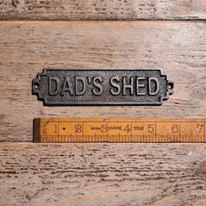 May include: A dark grey cast iron sign with the words "DAD'S SHED" in raised lettering. The sign is rectangular with decorative edges. A wooden ruler is below the sign.