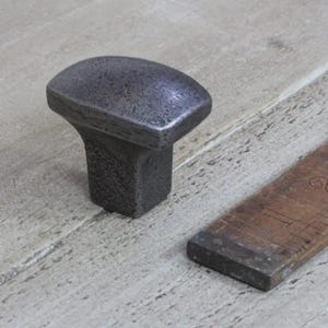 May include: A dark grey, textured metal cabinet knob with a rectangular base and a rounded top. A wooden ruler is partially visible in the background. The knob has a rustic, industrial aesthetic.