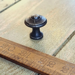 Peut inclure: Un bouton de meuble de style antique foncé avec un design en forme de champignon. Le bouton est en métal foncé avec une finition texturée. Une règle en bois avec des marquages en centimètres est au premier plan.