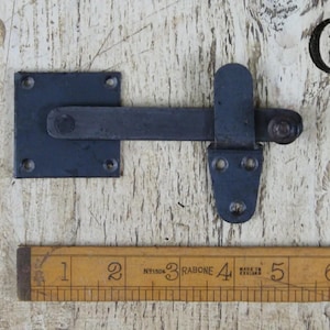 Op de afbeelding: Zwarte metalen deurgrendel met een vierkante plaat en een hendel. De grendel is ongeveer 10 cm lang.