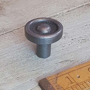 May include: A dark grey, round cabinet knob with a central raised circle and a short, cylindrical stem. The knob has a textured, aged appearance, suggesting a vintage or industrial style. The knob is likely made of metal.