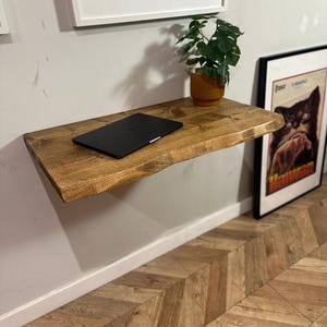 K&ouml;nnte beinhalten: Ein rustikales, schwebendes Holzregal mit einer nat&uuml;rlichen Holzoptik. Ein Laptop und eine Topfpflanze stehen auf dem Regal. Das Regal ist an einer wei&szlig;en Wand &uuml;ber einem Chevron-Muster-Holzfu&szlig;boden montiert.