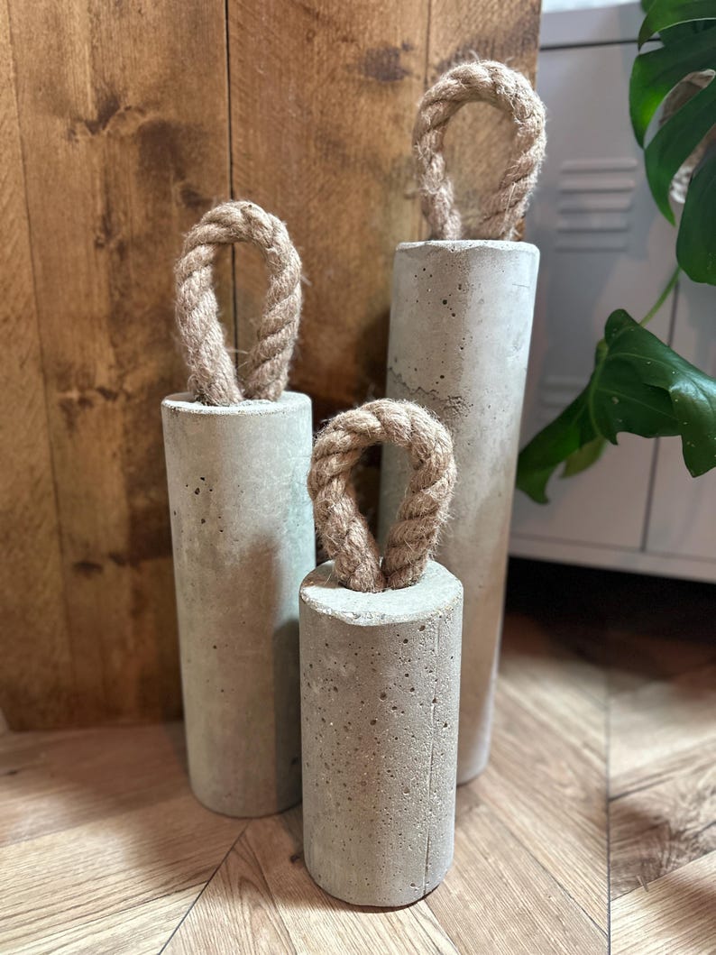 May include: Three concrete doorstops in different sizes with rope handles. The doorstops are a light grey colour and have a rough texture.