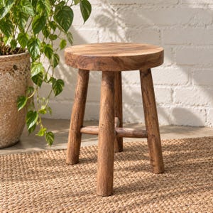 May include: A round wooden stool with a natural brown finish, featuring three sturdy cylindrical legs connected by a central cross-brace support structure. The stool has a smooth, flat circular seat and a visible wood grain texture throughout.