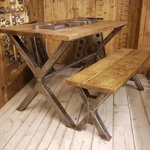 May include: A rustic wooden table with a metal X-shaped base and a matching bench. The table and bench are made of reclaimed wood and have a natural finish. The table is perfect for a dining room or kitchen.