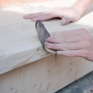 May include: A person sanding a piece of wood with a sanding block. The wood is light brown and the sanding block is dark brown.