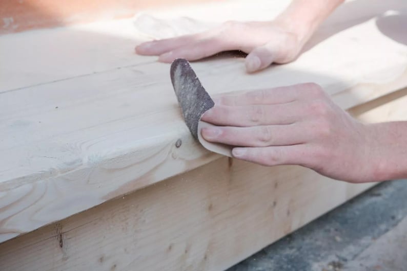May include: A person sanding a piece of wood with a sanding block. The wood is light brown and the sanding block is dark brown.