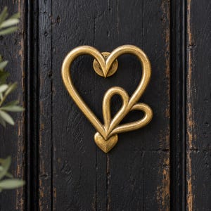 May include: A brass-toned metal door knocker shaped like two overlapping hearts. The design features a large heart outline with a smaller heart nested inside, both connected at the base. The finish has a slightly weathered, antique appearance.