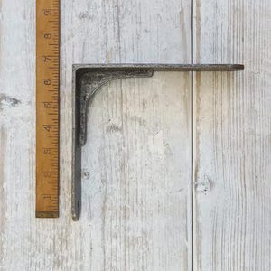 May include: A vintage-style, L-shaped metal shelf bracket with a distressed, gray finish. The bracket is shown next to a wooden ruler, with measurements in inches. The bracket is designed for supporting shelves and other items.