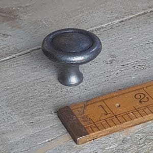 May include: A distressed, grey metal cabinet knob with a circular top and a short, cylindrical stem. The knob is on a grey wooden surface, with a vintage wooden ruler nearby.