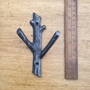 May include: A black metal wall hook shaped like a tree branch with two prongs. The hook is about 5 inches tall and is shown against a wooden background.