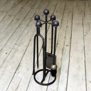 Puede incluir: Un juego de herramientas para chimenea de metal negro con un soporte. El juego incluye un atizador, unas pinzas, una pala y un cepillo. Las herramientas se mantienen en su lugar mediante un soporte circular con cinco bolas de metal negro.