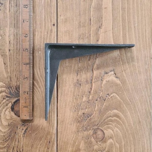May include: A black, L-shaped shelf bracket made of cast iron. The bracket is shown on a wooden surface next to a wooden ruler. The bracket is approximately 8 inches long.