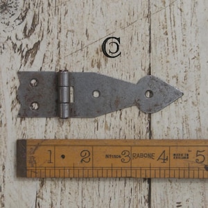 Può includere: Una cerniera in metallo rustico con un design decorativo a punta di freccia. La cerniera ha tre fori per viti e un perno centrale. Un righello di legno con misure in centimetri è sotto.