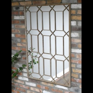 May include: Rectangular wall mirror with a geometric design. The mirror has a gold-coloured metal frame with a repeating pattern of rectangles and octagons. The mirror is mounted on a brick wall.