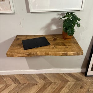 K&ouml;nnte beinhalten: Ein schwebendes Holzregal mit einem Laptop und einer Topfpflanze darauf. Das Regal ist an einer wei&szlig;en Wand &uuml;ber einem Holzboden mit Chevron-Muster befestigt.