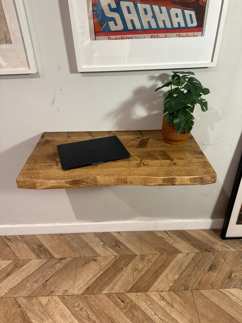 K&ouml;nnte beinhalten: Ein schwebendes Holzregal mit einem Laptop und einer Topfpflanze darauf. Das Regal ist an einer wei&szlig;en Wand &uuml;ber einem Holzboden mit Chevron-Muster befestigt.