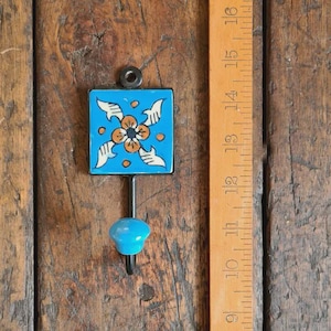 May include: A turquoise ceramic wall hook with a floral design in orange and white. The hook has a black metal stem and a turquoise knob. The hook is mounted on a wooden surface.