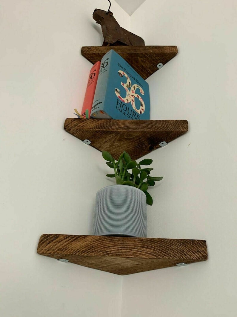 May include: Three wooden corner shelves with a rustic brown finish. The top shelf has a small metal animal figurine, two books, and the text "36 HOURS USA & CANADA". The middle shelf has a small potted plant. The bottom shelf is empty.