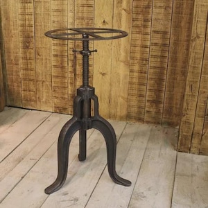 Op de afbeelding: Een verstelbare kruk van donker metaal met een ronde zitting en een driepoot. De zitting heeft een diameter van 35,5 cm. De kruk heeft een schroefdraad voor hoogteverstelling, tegen een achtergrond van houten planken.