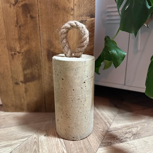 May include: A concrete doorstop with a rope handle. The doorstop is a light grey colour and has a rough texture. The rope handle is a light brown colour.
