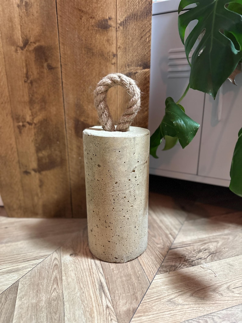 May include: A concrete doorstop with a rope handle. The doorstop is a light grey colour and has a rough texture. The rope handle is a light brown colour.
