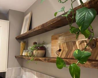 Op de afbeelding: Twee donkerbruine houten zwevende planken aan de muur. De bovenste plank bevat een ingelijste print. De onderste plank bevat een glazen pot met pasta, kleine kruidenpotjes, een tweekleurige keramische kom met een plant en twee snijplanken.