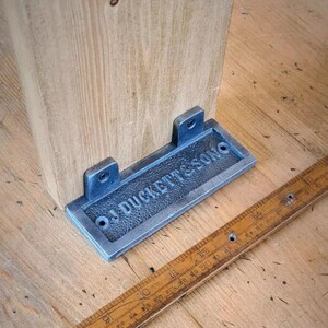 Puede incluir: Primer plano de un soporte de metal plateado con el texto "J. Duckett & Son" en relieve. El soporte está fijado a una viga de madera.