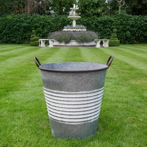Könnte beinhalten: Ein verzinkter Metalleimer mit zwei gebogenen Griffen. Der Eimer hat eine graue Oberfläche mit horizontalen weißen Streifen. Der Eimer steht auf einem grünen Rasen mit einem Brunnen und Hecken im Hintergrund.