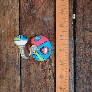 May include: A decorative wall hook with a colorful, hand-painted design. The hook features a ceramic knob with blue, green, and white accents, and a matching backplate with a floral pattern. The hook is approximately 3 inches long.