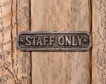 May include: A rectangular metal sign with a weathered dark gray finish featuring the embossed text STAFF ONLY in capital letters. The sign has beveled corners and is secured by two visible screws, one on each end of the horizontal plate.