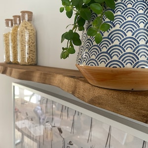 Puede incluir: Una estantería de madera con un borde natural, que presenta tres tarros de vidrio llenos de pasta y una maceta de cerámica azul y blanca con una planta verde. La estantería está montada en una pared blanca sobre una imagen enmarcada.