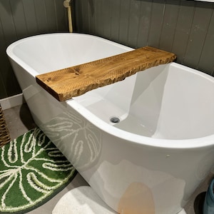 Peut inclure: Une baignoire blanche avec une étagère en bois fixée sur le côté. L'étagère est en bois naturel avec une finition rustique. La baignoire est posée sur un tapis de bain blanc avec un motif de feuille de monstera verte.