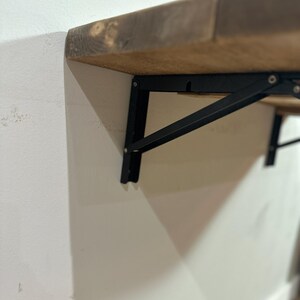K&ouml;nnte beinhalten: Ein Holzregal mit schwarzen Metallkonsolen, die an einer wei&szlig;en Wand befestigt sind. Das Regal ist aus Naturholz mit rustikalem Finish.