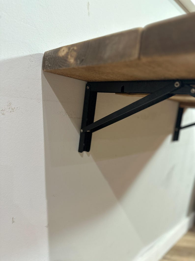 K&ouml;nnte beinhalten: Ein Holzregal mit schwarzen Metallkonsolen, die an einer wei&szlig;en Wand befestigt sind. Das Regal ist aus Naturholz mit rustikalem Finish.