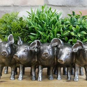 May include: A silver-coloured planter featuring five pig figurines, each with detailed features. The pigs are arranged side-by-side, holding a bed of green artificial plants. The planter is set on a wooden surface against a brick wall.
