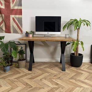 Op de afbeelding: Een houten bureau met zwarte metalen poten op een houten vloer met chevron-patroon. Een computermonitor, een potplant en een kopje staan op het bureau.