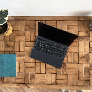 Op de afbeelding: Een houten tafelblad met een betegeld patroon. De tafel heeft een laptop, een blauw notitieboekje met de tekst "Tiffany & Co." op de omslag en een blauwe en witte keramische pot.