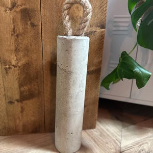 May include: A grey concrete doorstop with a rope handle. The doorstop is sitting on a wooden floor.