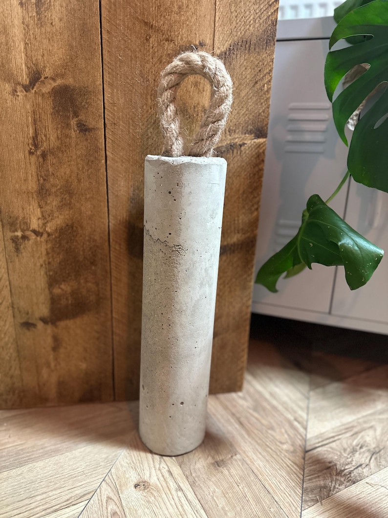 May include: A grey concrete doorstop with a rope handle. The doorstop is sitting on a wooden floor.