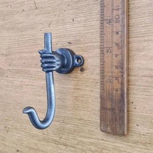 May include: A black cast iron hook shaped like a hand holding a curved rod. The hook is mounted on a wooden surface.